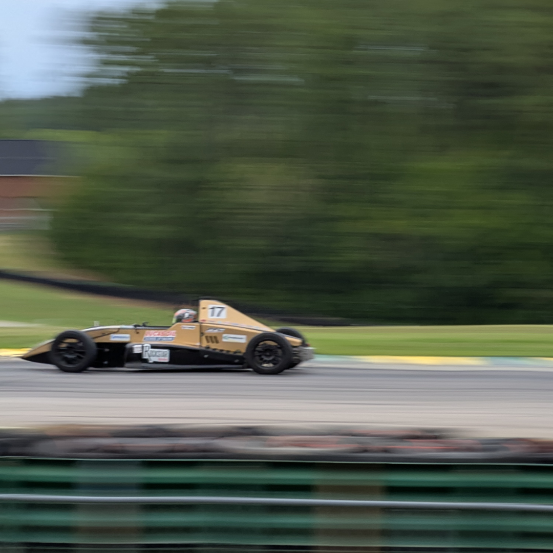 Brown vintage race car with number 17 speeding along a track, blurred green scenery in the background